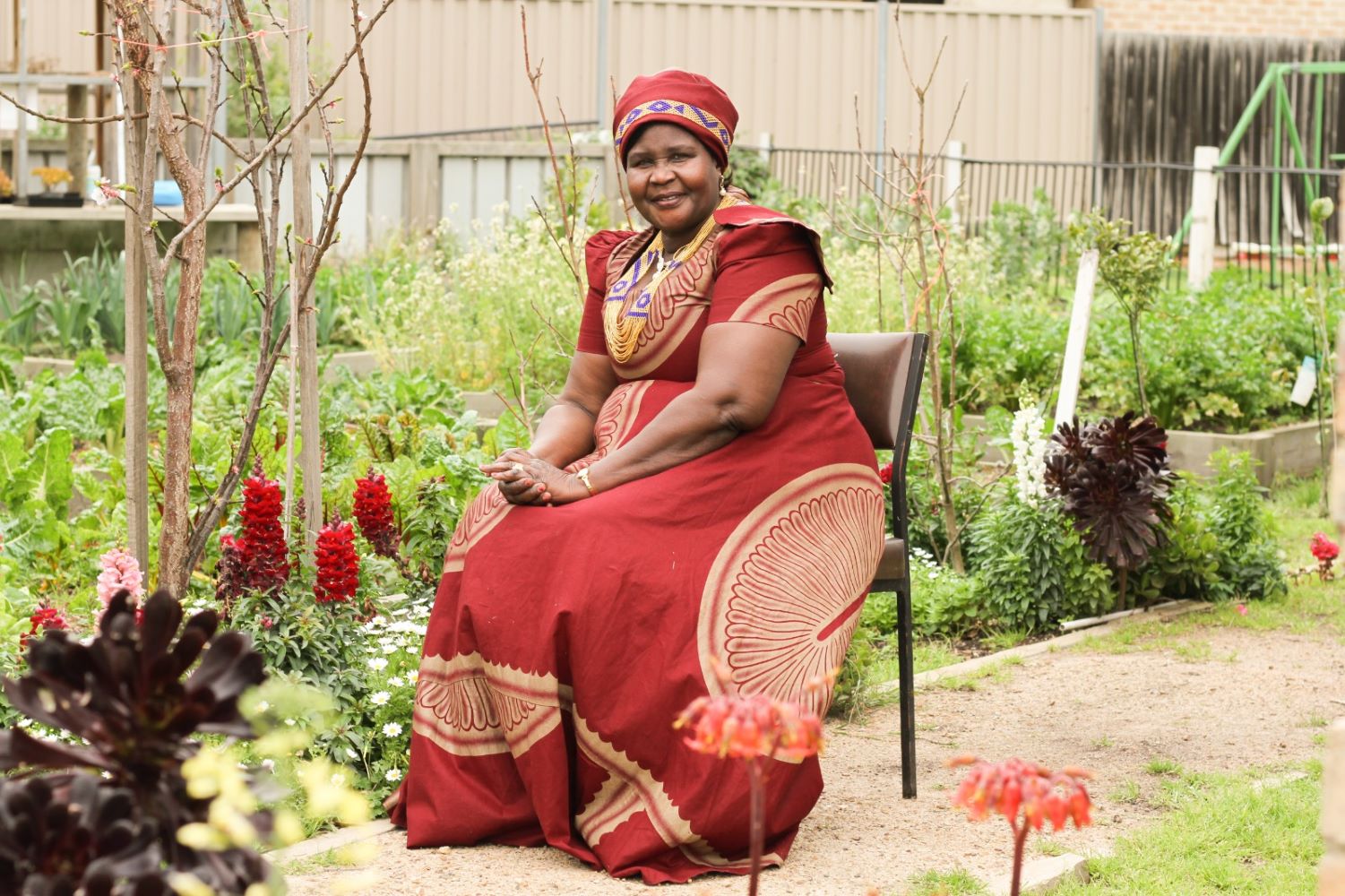 A smiling lady wearing traditional red African dress with head wear siting in a chair in a garden